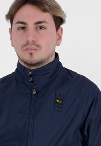 Giacca leggera blu navy con colletto alto, due bottoni e chiusura con zip. Presenta una tasca sul petto con etichetta logo.