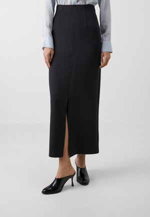 Woman wearing a high-waisted black midi skirt with front slit, light blue blouse, and black heeled mules, standing on a plain white floor.