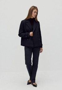 Marinblå dubbelknäppt sjömansrock med en strukturerad design, bärs över en svart polotröja, tillsammans med matchande mörka byxor och svarta flats.