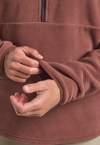 Haut en polaire de couleur brun-rouge, doté d'un col zippé mi-haut, d'une texture douce et de poignets élastiqués. Des mains ajustent les manches.