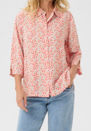 Vrouw draagt een lichtroze blouse met kleine rode en groene bloemenprint en blauwe spijkerbroek, één hand in de zak.
