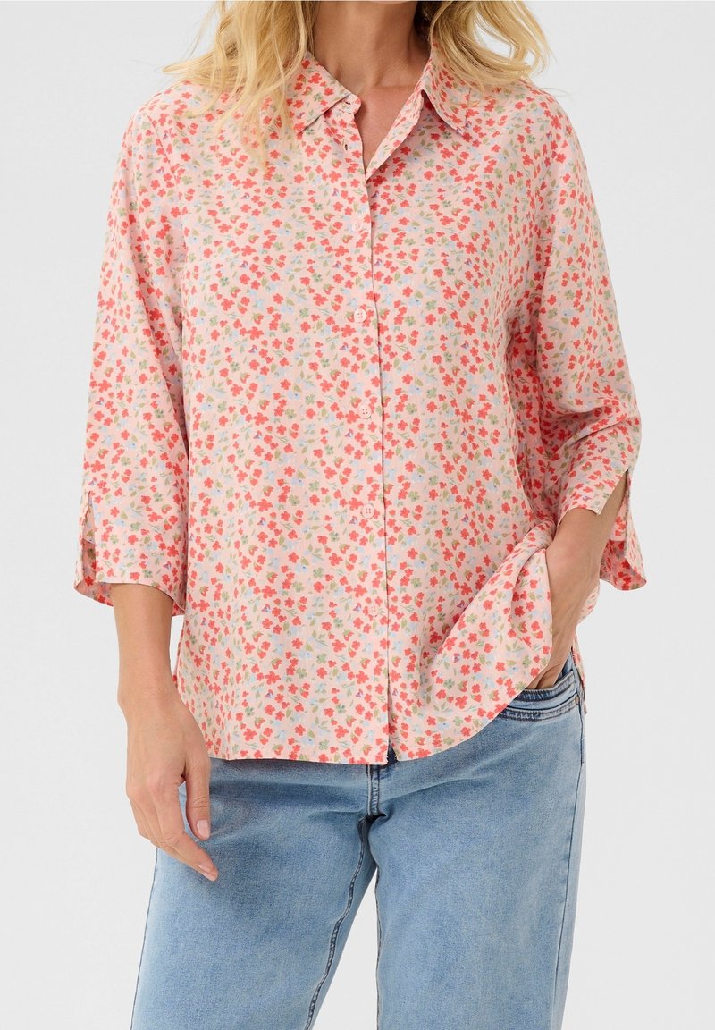 Femme portant une chemise rose pâle à boutons avec un petit imprimé floral rouge et vert et un jean bleu, une main dans la poche.