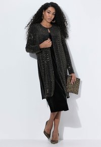 Svart paljettlång cardigan över en svart sammetklänning, kombinerad med en leopardmönstrad kuvertväska och metalliska högklackade skor. Texturerad, glamourös finish.