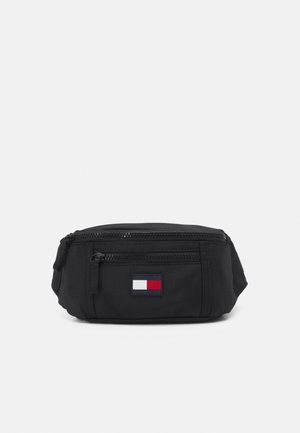 Zwarte stoffen heuptas met een ritsvak aan de voorkant en een klein rechthoekig logo met rood, wit en marineblauw.