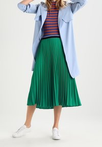 Abrigo oversized azul claro, blusa ajustada de rayas rojas y azules, falda midi plisada verde y zapatillas blancas. Tejido texturizado con pliegues definidos.
