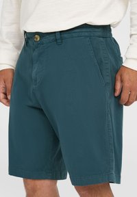 Pantaloni corti turchesi realizzati in un tessuto leggero, con una frontale piatta, tasche laterali e un dettaglio a bottoni in vita. Design casual con vestibilità comoda.