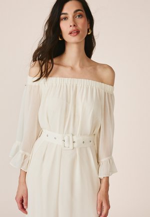 Woman wearing off-shoulder white dress with sheer long sleeves and a wide belt, standing against a neutral background.