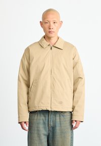 Bald man wearing beige zip-up jacket with large collar and blue faded jeans, standing against a plain white background.