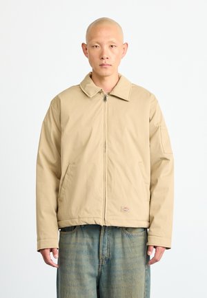 Bald man wearing beige zip-up jacket with large collar and blue faded jeans, standing against a plain white background.