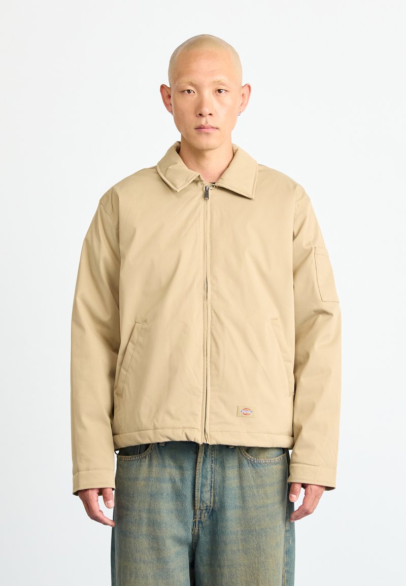 Bald man wearing beige zip-up jacket with large collar and blue faded jeans, standing against a plain white background.