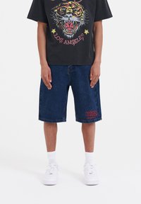 Denim shorts in dark blue with a relaxed fit, featuring red embroidered text on the left leg and a straight hem. Paired with a black graphic tee.