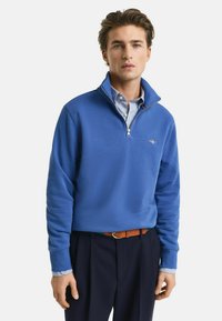 Royalblauer Pullover mit einem Reißverschlusskragen, aus weichem Stoff gefertigt. Verfügt über einen gemusterten Innenkragen und ein kleines gesticktes Logo auf der Brust.