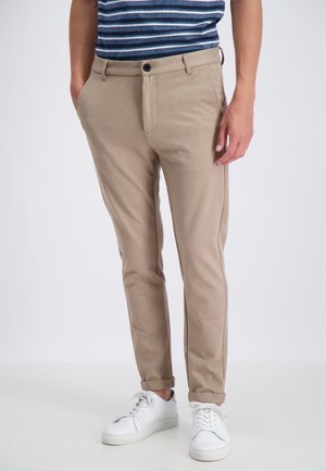 Mężczyzna ubrany w beżowe spodnie slim fit, białe sneakersy oraz niebieską koszulę w paski, stojący z rękami w kieszeniach na jednolitym tle.