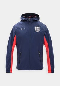 Σκούρο μπλε μπουφάν με κουκούλα και κόκκινα πλαϊνά πάνελ, τσέπες με φερμουάρ, το έμβλημα της ποδοσφαιρικής ομάδας της Αγγλίας με ένα αστέρι και λευκό λογότυπο Nike στο στήθος.