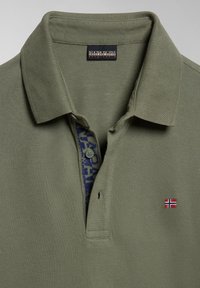 Polo shirt verde oliva con tessuto strutturato, colletto con bottoni e un interno della patta blu decorato. Presenta un piccolo emblema a forma di bandiera sul petto.