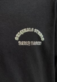 Melna kokvilnas T-krekls ar izliektu izšūtu logo gaiši pelēkā un baltā krāsā, kas lasāms "Originals Studio Daily Basics."