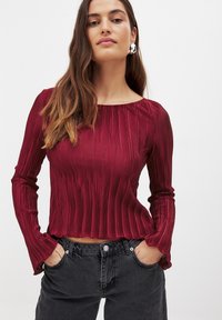 Rød plisseret langærmet top med rund halsudskæring, struktureret stof og flagrende manchetter, parret med mørke jeans med knapdetalje.
