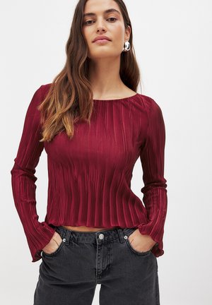 Red pleated long-sleeve top with a round neckline, textured fabric, and flared cuffs, paired with dark jeans featuring a button detail.