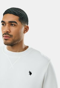 Felpa bianca con scollo rotondo, dettagli a coste e piccolo logo nero sul petto. Tessuto liscio e design minimalista.