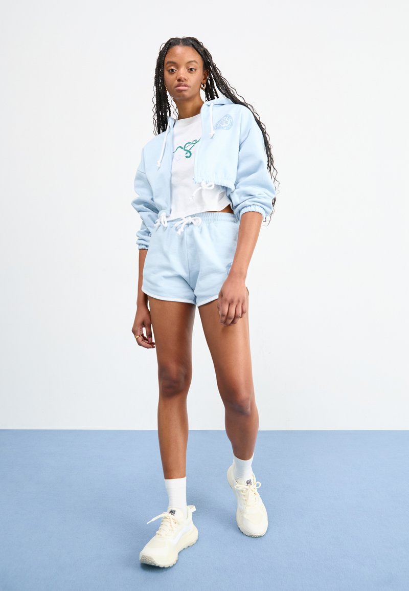 Chaqueta corta azul claro con cordón y logotipo, combinada con pantalones cortos a juego. Camiseta blanca por debajo, zapatillas color crema y calcetines blancos.