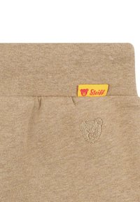 Tela de algodón beige con una textura suave, que presenta un pequeño logo de oso bordado y una etiqueta amarilla con texto rojo.