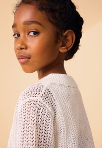 Jauna mergina su tamsiais garbanotais plaukais ir baltu megztiniu, žvelgianti per petį prieš smėlio spalvos foną.