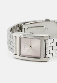 Reloj de acero inoxidable plateado en forma de cuadrado con una esfera de color rosa pálido y movimiento de tres agujas. Presenta una pulsera de malla metálica y un detalle de logo.