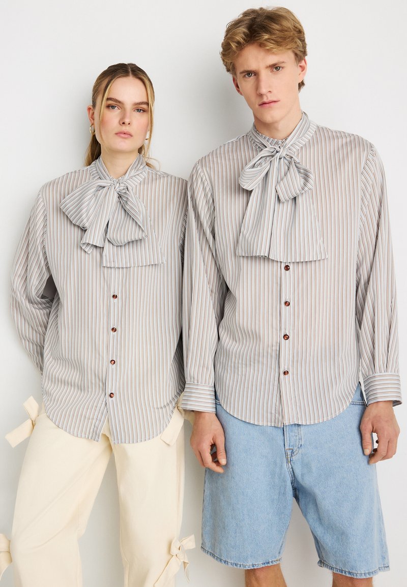 Ριγέ πουκάμισα από ελαφρύ ύφασμα, με παπιγιόν στον γιακά και καφέ κουμπιά. Ένα πουκάμισο έχει κάθετες μπεζ και καφέ ρίγες.