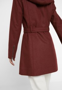 Bordový vlněný kabát s kapucí, páskem v pase a zipy na rukávech. Vyznačuje se přizpůsobeným střihem a měkkou, strukturovanou tkaninou.