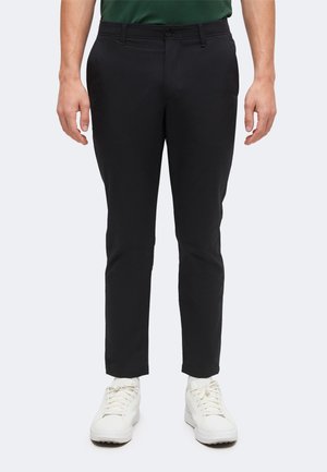 Homme portant un pantalon noir slim, une chemise verte rentrée et des baskets blanches, debout devant un fond clair uni.