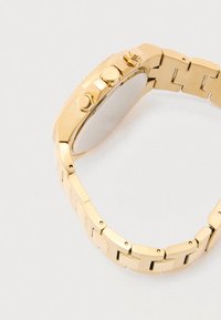 Zlata kovinska ura z obliko zapestnice iz povezav, okroglo ohišje, tremi gumbi na strani in gladko, polirano površino.