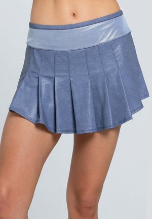 Vrouw draagt een korte, geplooide, glanzende blauwe sportrok met een brede tailleband tegen een effen lichtgrijze achtergrond.
