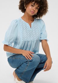 Világoskék blúz rövid puffos ujjal, V-nyakkivágással és hímzett cseppmintával. Kékfarmer csípős nadrággal és világos lábbelivel párosítva.