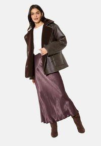 Pruun nahktagi fliisist kraega, valge kampsun, lilla satiinist seelik ja pruunidest seemisnahast põlvikud. Modell seisab neutraalsel taustal.