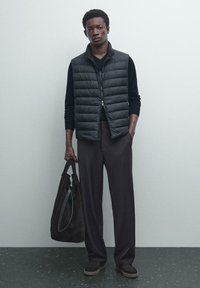 Homme portant un gilet matelassé noir sur une chemise à manches longues noire, un pantalon large sombre, tenant un grand sac fourre-tout noir, se tenant contre un mur clair.