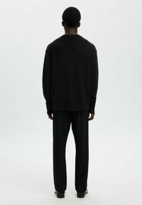 Hombre con cabello corto y rizado, vistiendo un suéter negro, pantalones negros y zapatos negros, de pie frente a un fondo blanco liso.