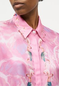 Približni prikaz osobe u ružičastoj košulji s gumbima, šiljastim ovratnikom i ukrasnim cvjetnim uzorcima u nježnim bojama.