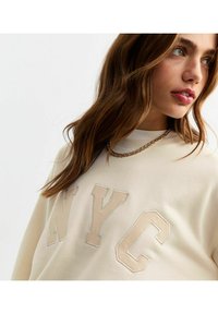 Krämfärgad sweatshirt med ett pr präglat "NYC"-logotyp i matchande tyg, med ribbad halsringning och strukturerad yta. Guld kedja synlig.