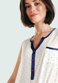 Top sin mangas de tela color crema con pequeños lunares rojos y azules, cuello navy, tapeta de botones y un único bolsillo en el pecho.