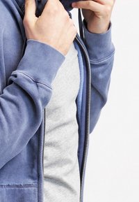 Felpa con cappuccio blu con cerniera, tasche, tessuto morbido, polsini a costine e cordoncini, indossata sopra una t-shirt grigia chiara, che mostra strati a contrasto.