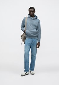 Grå fleecetrøje med kængurulomme, lyseblå denimjeans og hvide sneakers med sorte detaljer. Modellen bærer en beige taske.