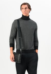 Suéter gris de espiga con cuello alto negro, combinado con un pequeño bolso de cuero negro tipo bandolera. Unos pantalones negros sólidos completan el look.