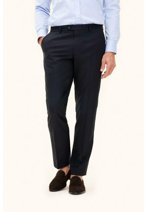 Uomo che indossa pantaloni formali neri, camicia azzurro chiaro e mocassini in pelle scamosciata marrone scuro con le mani nelle tasche su sfondo semplice.