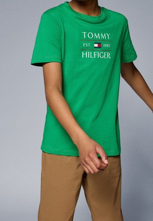 Person iført en grøn Tommy Hilfiger T-shirt med logo og beige bukser, mod en ensfarvet grå baggrund.