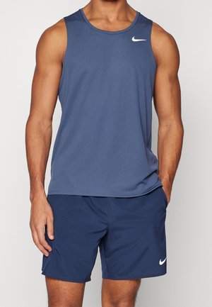 Muskeløs person iført en blå ærmeløs sportstop og matchende blå shorts med hvide Nike-logoer, stående med afslappede hænder.