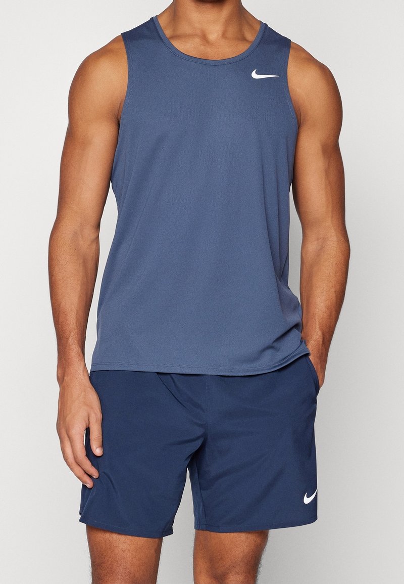 Personne musclée portant un haut de sport bleu sans manches et un short bleu assorti avec des logos blancs Nike, debout les mains détendues.