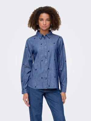 Femme aux cheveux bouclés portant une chemise en jean bleu à boutons avec de petits motifs de cœurs foncés et un jean bleu assorti, debout.