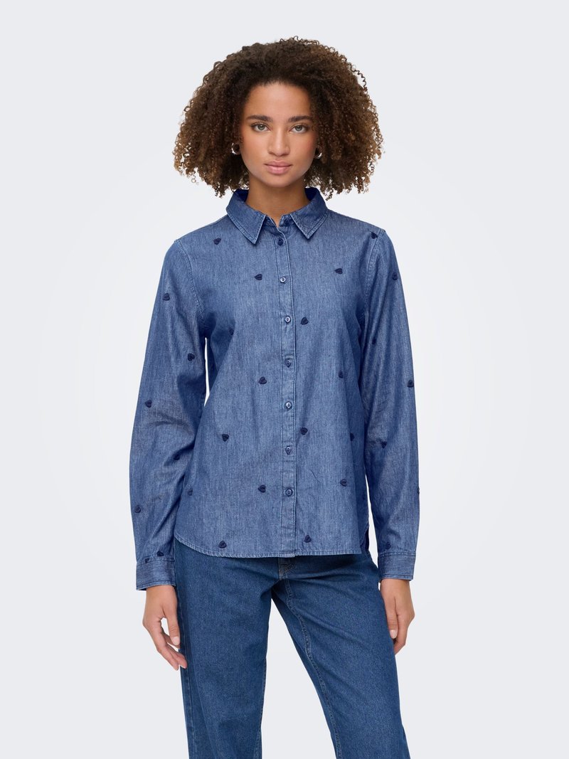 Femme aux cheveux bouclés portant une chemise en jean bleu à boutons avec de petits motifs de cœurs foncés et un jean bleu assorti, debout.