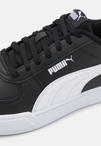 Musta synteettinen lenkkari, jossa on valkoinen PUMA-logo ja korostus. Vaatimuksena on teksturoitu päällinen, pyöreä varvas ja litteä valkoinen kumipohja.