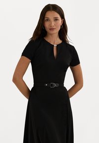 Μαύρο φόρεμα με κοντά μανίκια, λαιμόκοψη σε σχήμα κλειδώματος και ζωνάκι στη μέση. Διαθέτει λεπτή ζώνη με στρογγυλή αγκράφα και απαλό υφασματικό υλικό.